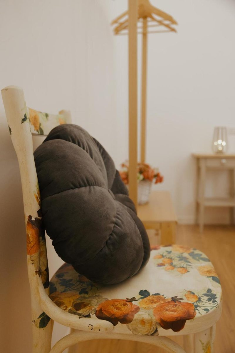 Sedia in legno curvato con la seduta decorata a fiori e due cuscini grigi arrotolati, accanto all’appendiabiti in legno